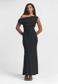 Mustat off-the-shoulder-mekko, jossa on valumateriaali, tiukka siluetti ja lattiaan asti ulottuva muotoilu. Liukas pinta ja minimalistinen tyyli.