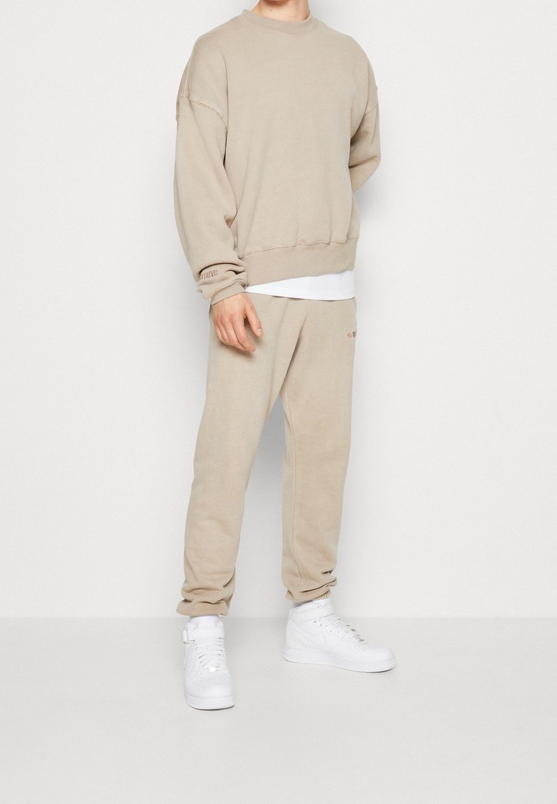 Homme portant un sweat-shirt beige et un jogging assorti avec des baskets blanches, debout devant un fond blanc uni.