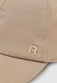 Béžová baseballová čepice s vyšitým písmenem „R“ na předním panelu, s viditelným šitím a texturovanou látkou.