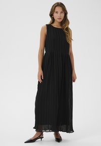 Svart, ärmlös, golvlång klänning med vertikala veck, rundad halsringning och rynkad midja. Bärs med spetsiga svarta skor.