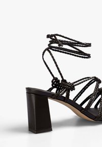 Svart klacksandals med flätade repremmar, en fyrkantig klack och en slät innersula. Designen framhäver intrikata knutdetaljer.