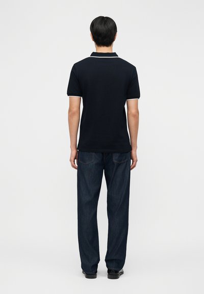 Άνδρας που στέκεται με την πλάτη, φοράει μαύρο πόλο με λευκή λεπτομέρεια και σκούρο μπλε τζιν, μπροστά σε σκηνικό με απλό φόντο.
