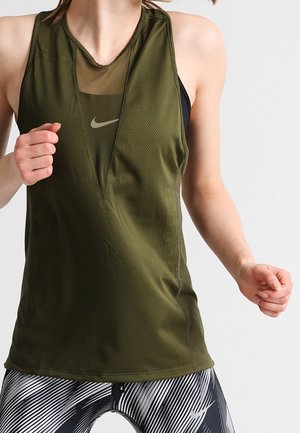 Femme portant un haut de sport sans manches vert olive avec un détail en mesh en V profond sur un soutien-gorge de sport noir, assorti à un legging noir et blanc à motifs.