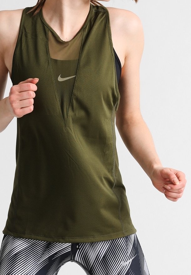 Femme portant un haut de sport sans manches vert olive avec un détail en mesh en V profond sur un soutien-gorge de sport noir, assorti à un legging noir et blanc à motifs.