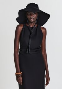 Schwarzes Neckholderkleid mit fließendem Rock, begleitet von einem schwarzen Hut mit breiter Krempe. Ausgestattet mit einem gebundenen Band und Holzarmreifen.