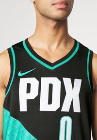 Maillot de basket noir avec des accents turquoise et "PDX" en grandes lettres blanches. Il possède un design en V et une texture en mesh, avec le logo Nike.