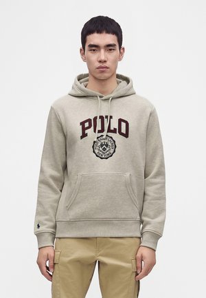 Młody mężczyzna w beżowej bluzie z kapturem Polo Ralph Lauren i khaki spodniach typu cargo stoi na jednolitym jasnym tle z neutralnym wyrazem twarzy.