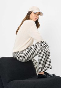 Lichtcrème, donzige trui; zwart-wit houndstooth-patroon broek; beige pet met blauwe accenten; zwarte instappers op een donkere, gestoffeerde stoel.