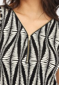 Černý látkový top s bílým vzorem kapradí, se zlatým zipem na výstřihu, který zdůrazňuje kontrast a texturu.