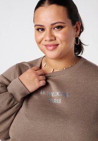 Bruine sweatshirt met glinsterende tekst "LE WEEKEND PARIS," geribde manchetten en zachte, textuurstof. Model houdt de kraag voorzichtig vast.