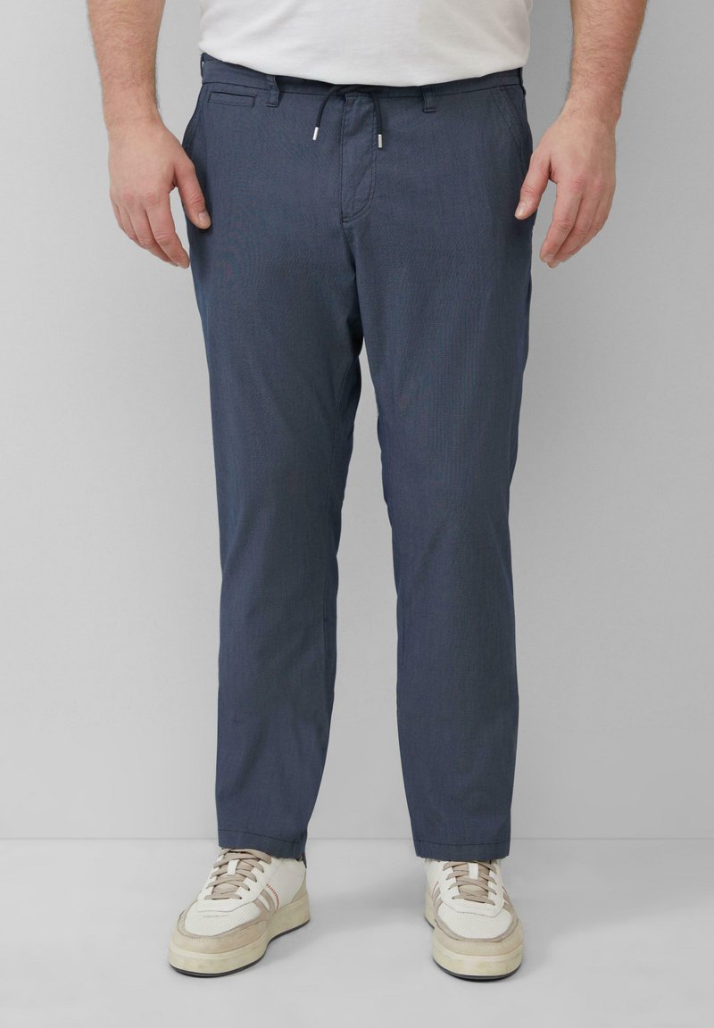 Navyblauwe, licht getextureerde broek met een trekkoord in de taille, zijzakken en een taps toelopende pijp, gecombineerd met witte en beige sneakers.