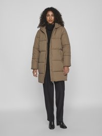 Vadderad brun parka med huva, med dragkedjestängning och stora quiltade sektioner. Buren över en svart rullkrage och svarta byxor.