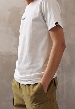 Personne portant un t-shirt blanc à manches courtes avec un petit logo sur la manche et un pantalon cargo vert olive, les mains dans les poches.