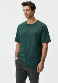 Ciemnozielona bawełniana koszulka z krótkimi rękawami. Posiada zaokrąglony dekolt i małe wytłaczane logo na piersi. Noszona z szarymi dżinsami.