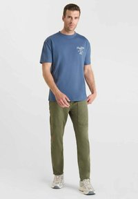 Hombre de pie con una camiseta gráfica azul, pantalones verdes oliva con parches en las rodillas y zapatillas blancas, frente a un fondo claro y liso.