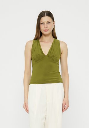 Γυναίκα με ίσια καστανά μαλλιά που φοράει αμάνικο τοπ σε ελιά πράσινο με V-λαιμόκοψη και λευκά ψηλόμεσα παντελόνια, στέκεται μπροστά σε απλό φόντο.