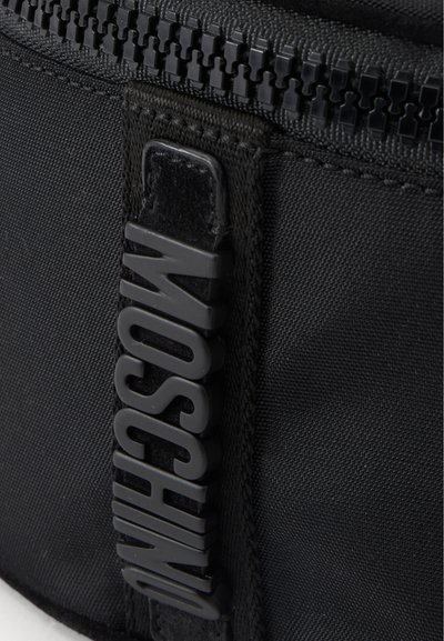 Gros plan d'un sac en tissu noir avec une fermeture éclair détaillée et une étiquette verticale avec des lettres en relief formant "MOSCHINO."