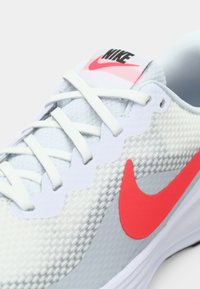 Šviesiai pilkos spalvos Nike sportiniai batai su tekstūruotu sintetine viršutinė dalimi, baltais raišteliais ir ryškiai raudonu swoosh. Turinti paminkštintą padą ir paminkštintą kraštą.
