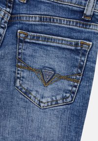 Zadní kapsa modrých džínsů s žlutým švem a trojúhelníkovou nášivkou loga Guess přišitou na střed kapsy.