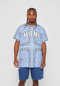 Blå grafisk t-skjorte med paisley-mønster, med hvite detaljer og ordet "KANI" i fet skrift. Brukt med denimshorts.