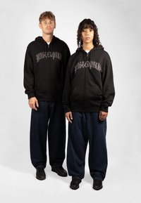 Schwarze Zip-Hoodies mit einem grafischen "Mastemind"-Druck. Kombiniert mit lockeren, dunkelblauen Jeans und schwarzen Sneakers. Glatte Textur.