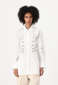 Camicia bianca con bottoni, caratterizzata da intricati motivi a taglio sulle maniche, realizzata in tessuto leggero. Presenta un colletto classico e maniche lunghe.