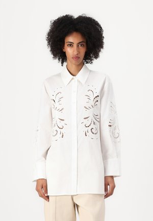 Camicia bianca con bottoni, caratterizzata da intricati motivi a taglio sulle maniche, realizzata in tessuto leggero. Presenta un colletto classico e maniche lunghe.