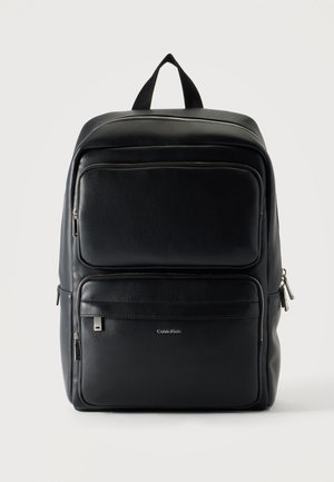 Musta nahkainen Calvin Klein -reppu, jossa useita vetoketjullisia lokeroita ja yläkantokahva, yksiväristä taustaa vasten.