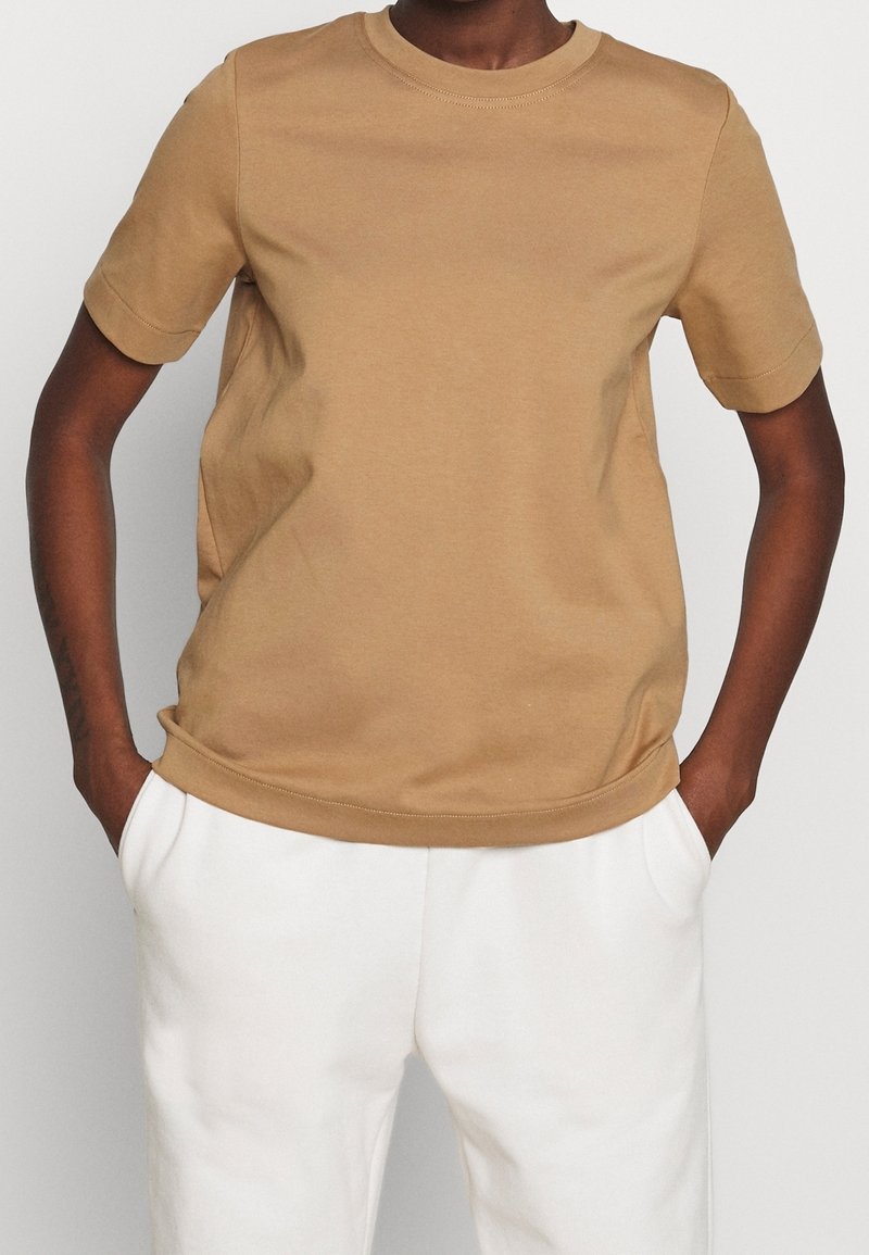Personne portant un t-shirt beige à manches courtes et un pantalon blanc, debout les mains dans les poches sur un fond uni.
