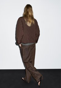 Person mit langen Haaren, der eine braune Steppjacke, lockere braune Hosen und schwarze Sandalen trägt, geht von einem weiß-schwarzen Hintergrund weg.