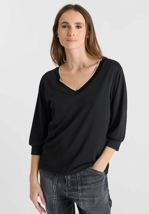 Femme portant un haut noir à col en V avec des bordures dorées et des manches trois-quarts, associé à un jean gris foncé, se tenant avec une main dans la poche.