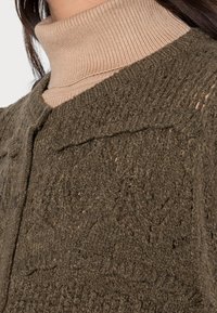 Cardigan vert olive en tricot avec un motif texturé, doté d'un col rond et d'une fermeture à boutons. Un col roulé beige est porté en dessous.