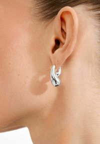 Boucle d'oreille argentée de forme abstraite avec une finition polie, présentant une texture lisse et un design incurvé, portée à l'oreille.