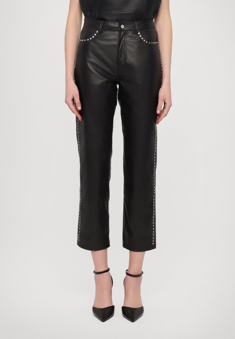 Pantalon en cuir noir court avec des clous argentés le long des poches et des coutures latérales, porté avec des talons noirs à bride cheville et bout pointu.