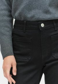 Mustat korkeavyötäröiset farkut, joissa on klassinen viisi-taskuinen muotoilu, hopeinen nappikiinnitys ja sileä, teksturoidun tuntuinen kangas.