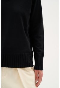Maglione nero a maniche lunghe con polsini e orlo a costine indossato con pantaloni beige chiaro, con focus su torso e mano destra.