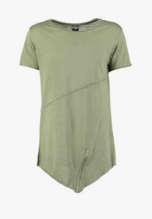 T-shirt à manches courtes, de couleur olive, avec un ourlet asymétrique et une couture diagonale. Comprend un détail zip sur le bas à droite.