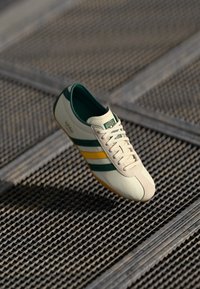 Tenis bej cu dungi verzi și galbene, material din piele, textură netedă, design cu șireturi și talpă din cauciuc, suspendat deasupra unei suprafețe în plasă.