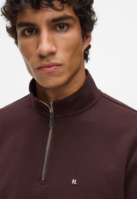 Burgundy színű, cipzáros pulóver magas gallérral, texturált anyagból és ezüst színű fémalkatrészekkel. A mellkasán kisebb logó található.