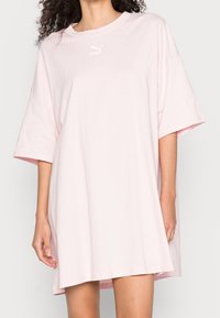 Różowa, bawełniana, oversize sukienka t-shirtowa z krótkimi rękawami, okrągłym dekoltem i małym białym logo na piersi. Gładka faktura, długość do kolan.