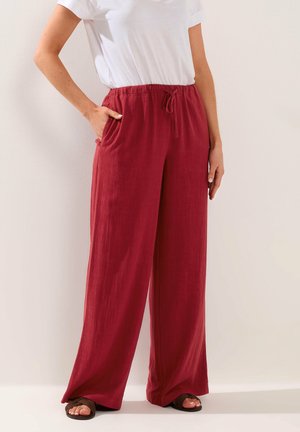 Osoba ubrana w luźne czerwone spodnie ze sznurkiem i białą koszulę z krótkim rękawem, z jedną ręką w kieszeni, stojąca na tle jednolitego jasnego tła.