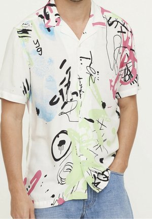 Man wearing short-sleeve white button-up shirt with colorful abstract graffiti designs and blue jeans, standing with one hand in pocket.