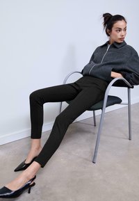 Pantaloni neri aderenti con una texture liscia, abbinati a una giacca grigia con zip. Il modello è seduto su una sedia, indossando tacchi a punta neri.