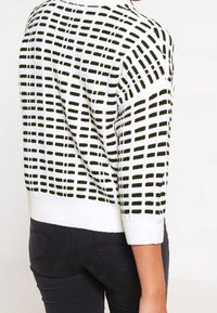 Vit tröja med ett svart rutmönster, rundad halsringning och trekvartslånga ärmar, med ribbad fåll och ärmslut.