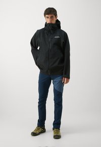 Jachetă impermeabilă neagră cu fermoar, buzunare laterale și guler ridicat. Asortată cu pantaloni bleumarin cu un accent albastru și pantofi de drumeție verzi.