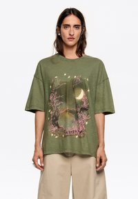 Olijfgroene oversized t-shirt met korte mouwen, voorzien van een grafische print van een maanlichtscène, takken en vogels in het roze.