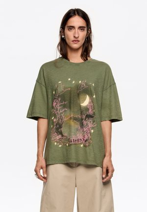 Camiseta oversize verde olivo con mangas cortas que presenta una impresión gráfica de una escena iluminada por la luna, ramas de árbol y pájaros en color rosa.