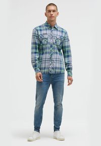 Herren-Karottenhemd in Blau, Grün und Weiß. Baumwollmaterial mit langen Ärmeln und Button-Down-Kragen. Zwei Brusttaschen, Slim Fit Jeans, weiße Sneaker.