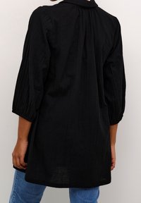 Personne portant un blouse noire à manches longues amples avec des poignets froncés, associée à un jean bleu, montrée de dos contre un mur uni.