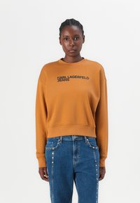 Bronzen sweatshirt met ronde hals en zwarte "KARL LAGERFELD JEANS" print. Gecombineerd met blauwe jeans met decoratieve knopen aan de naden.